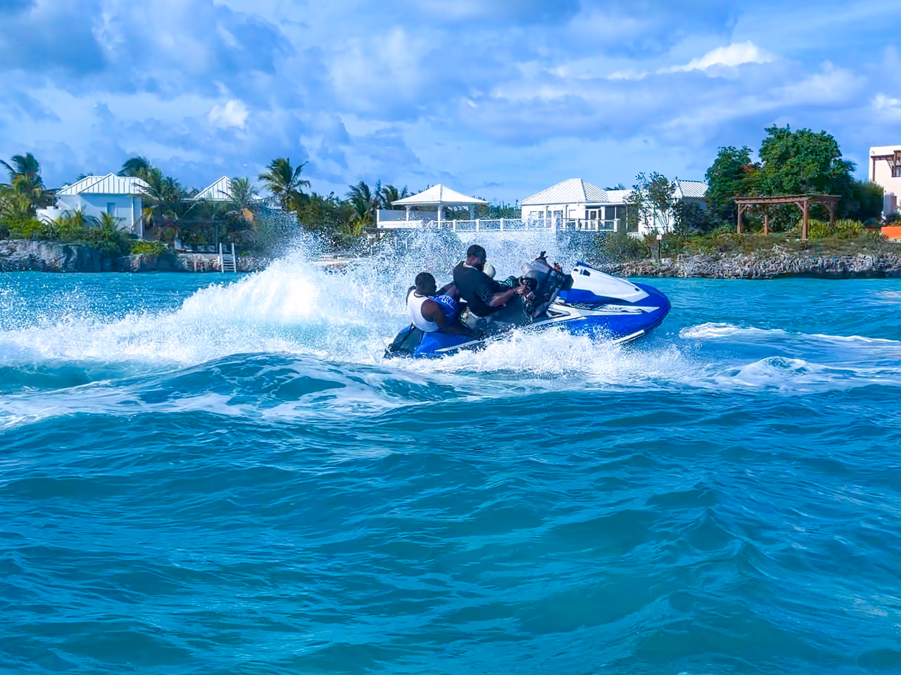 A blue and white Yamaha WaveRunner underway with two riders, spray behind it and white villas on the shore