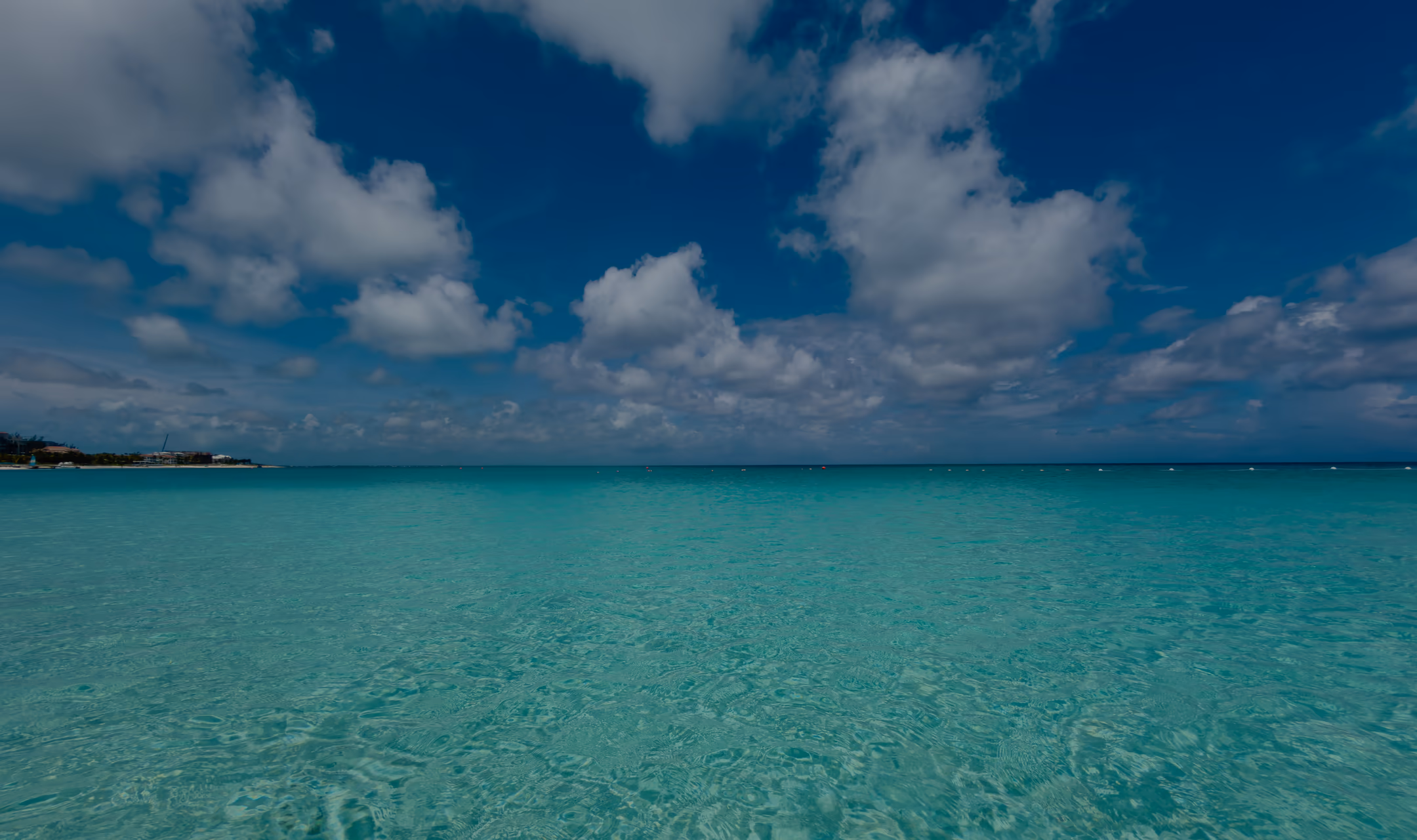Clear, shallow turquoise water at eye level, running out to a flat horizon under a deep blue sky and white clouds
