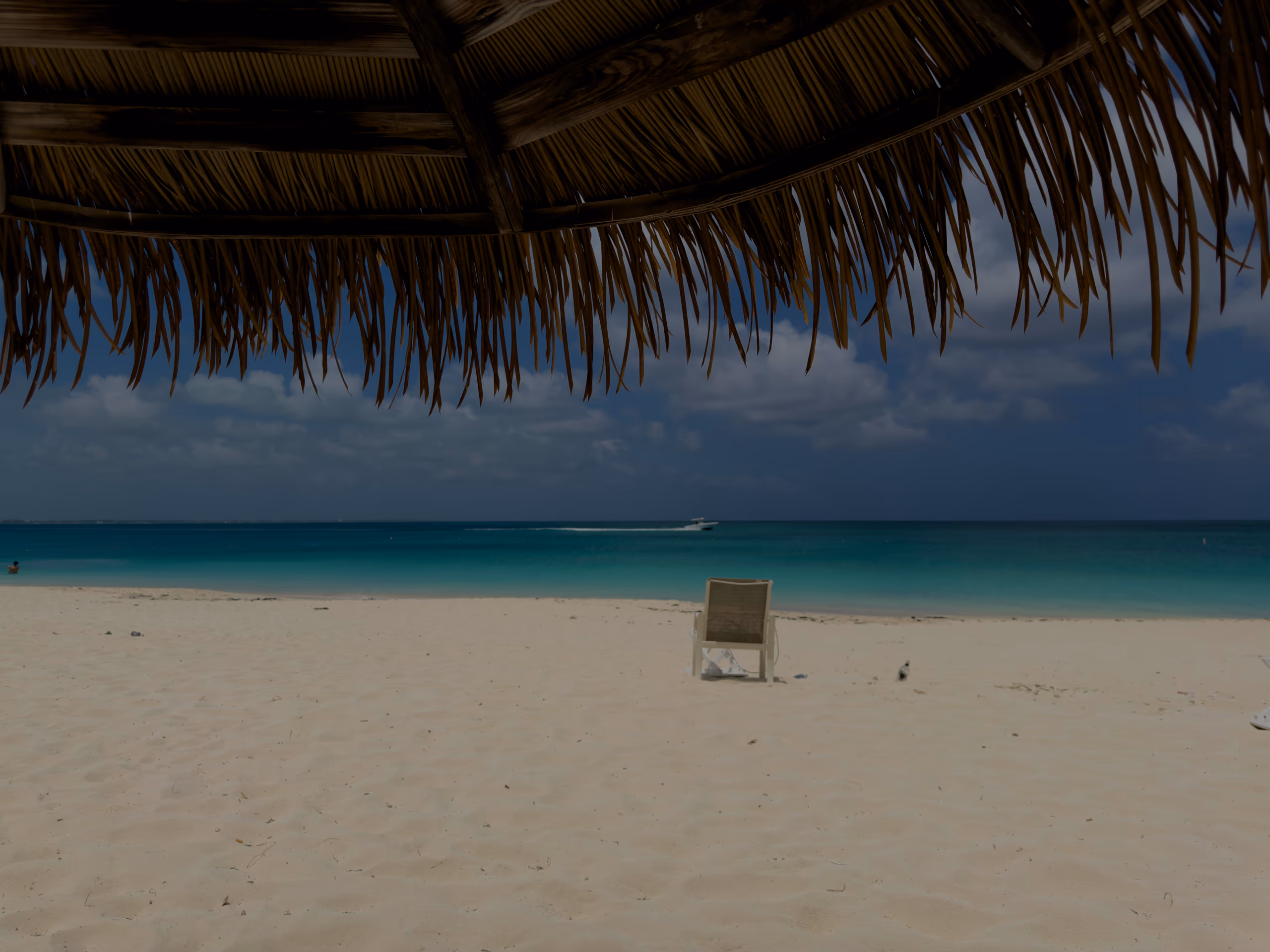 View from under a palapa across white sand to turquoise water, a boat on the horizon