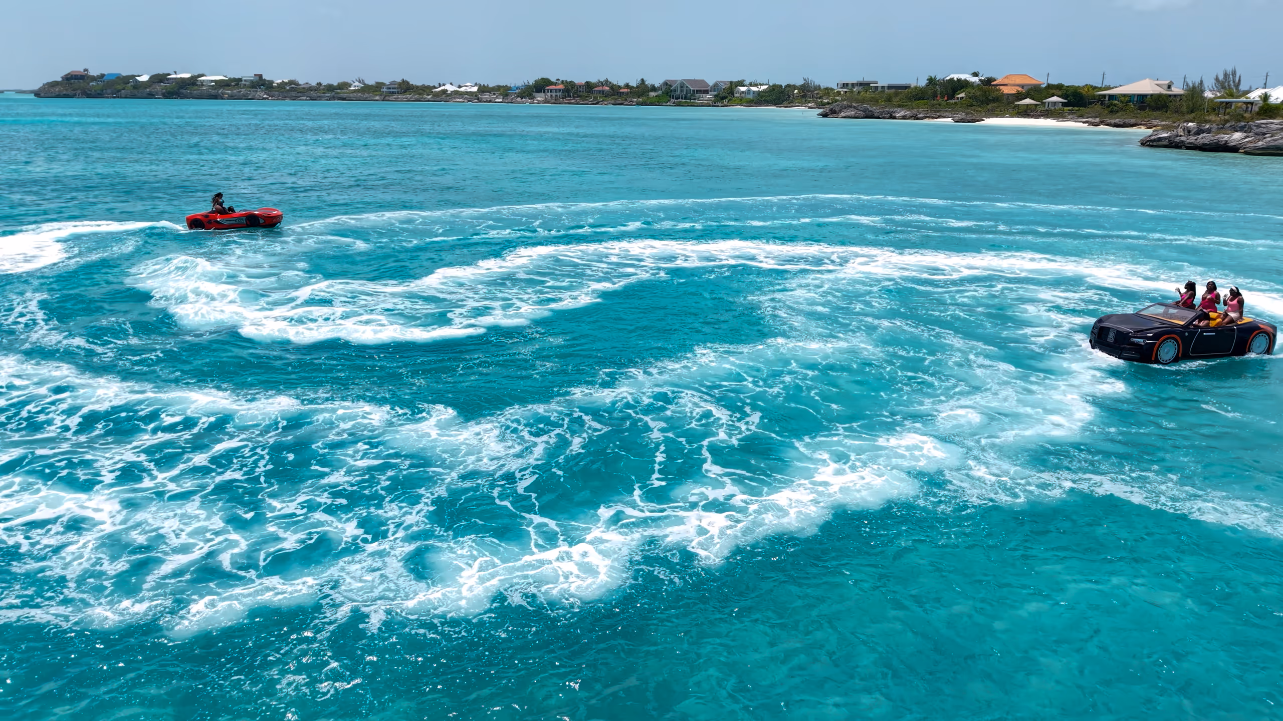 Both Jet Cars trailing white wakes across turquoise water, with the coastline behind, seen from our drone