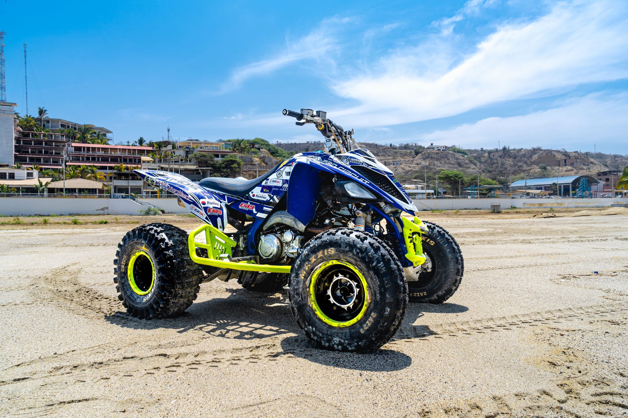 Blue and yellow sport quad parked on packed sand, a scrubby hillside behind it