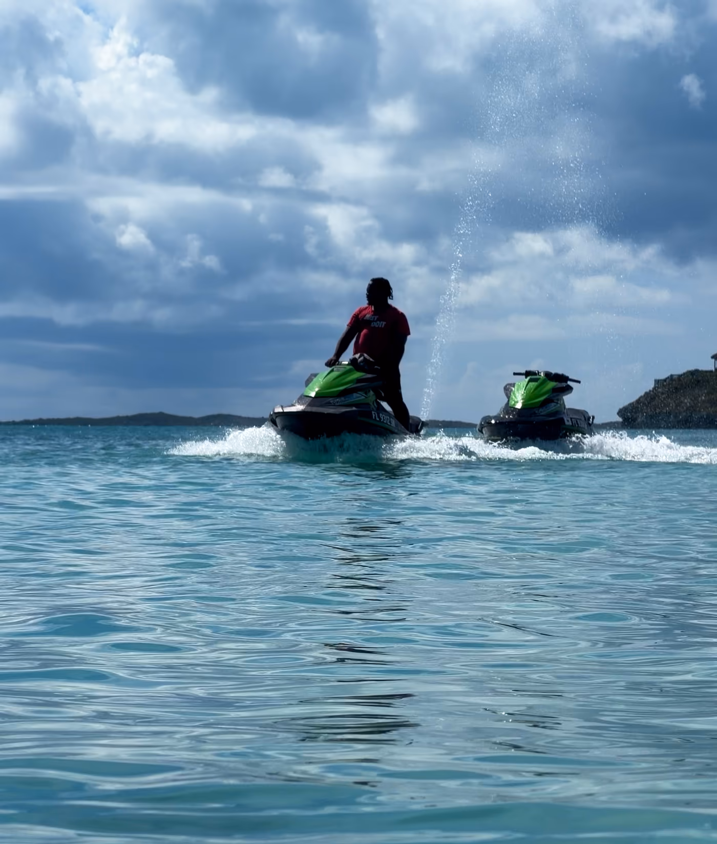 A rider heading out on a green and black Jet Ski across turquoise water, a second Jet Ski beside them and a rocky point on the right