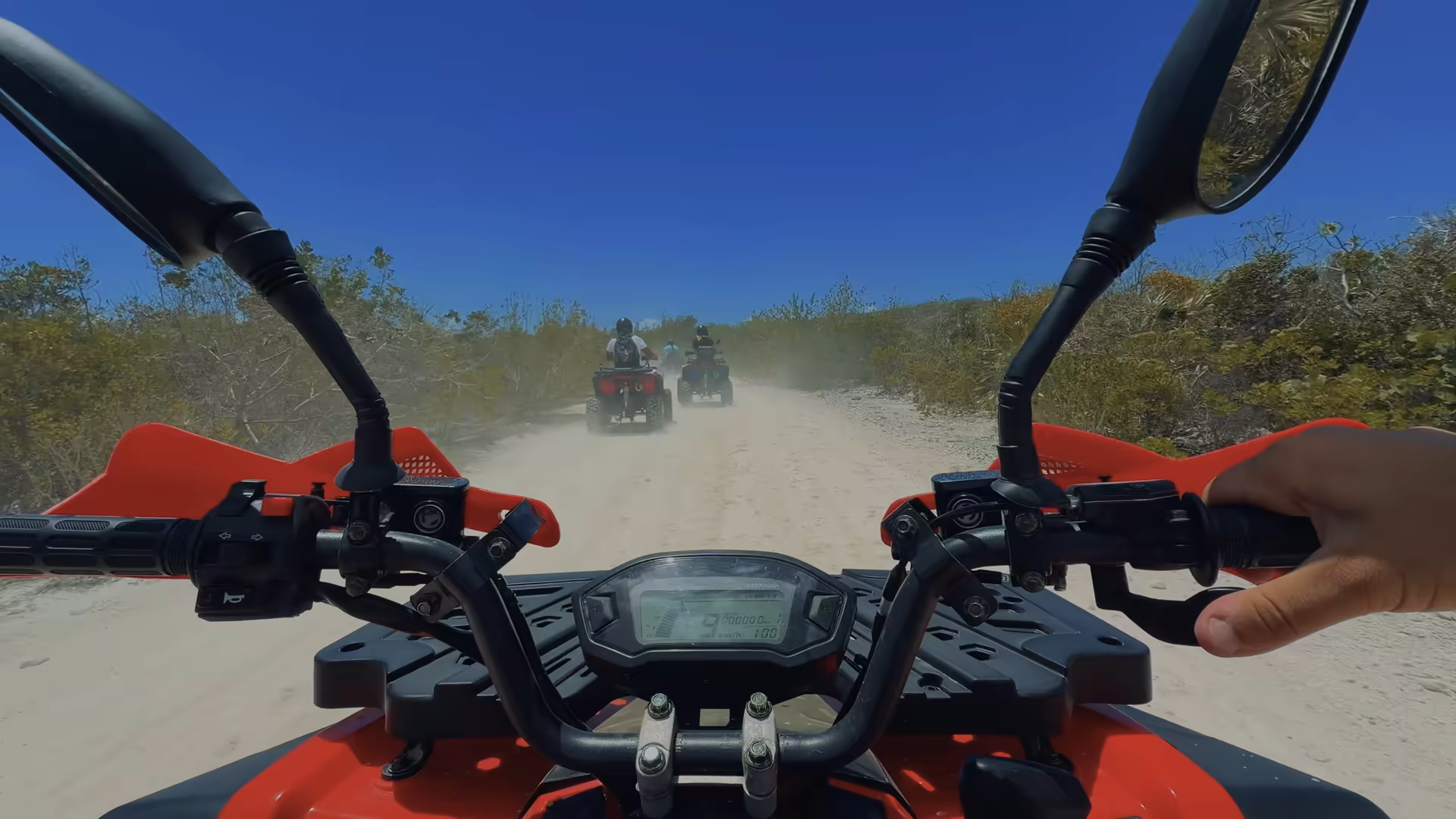 A rider's view over red ATV handlebars, following a line of quads down a dusty white track through the scrub of Providenciales