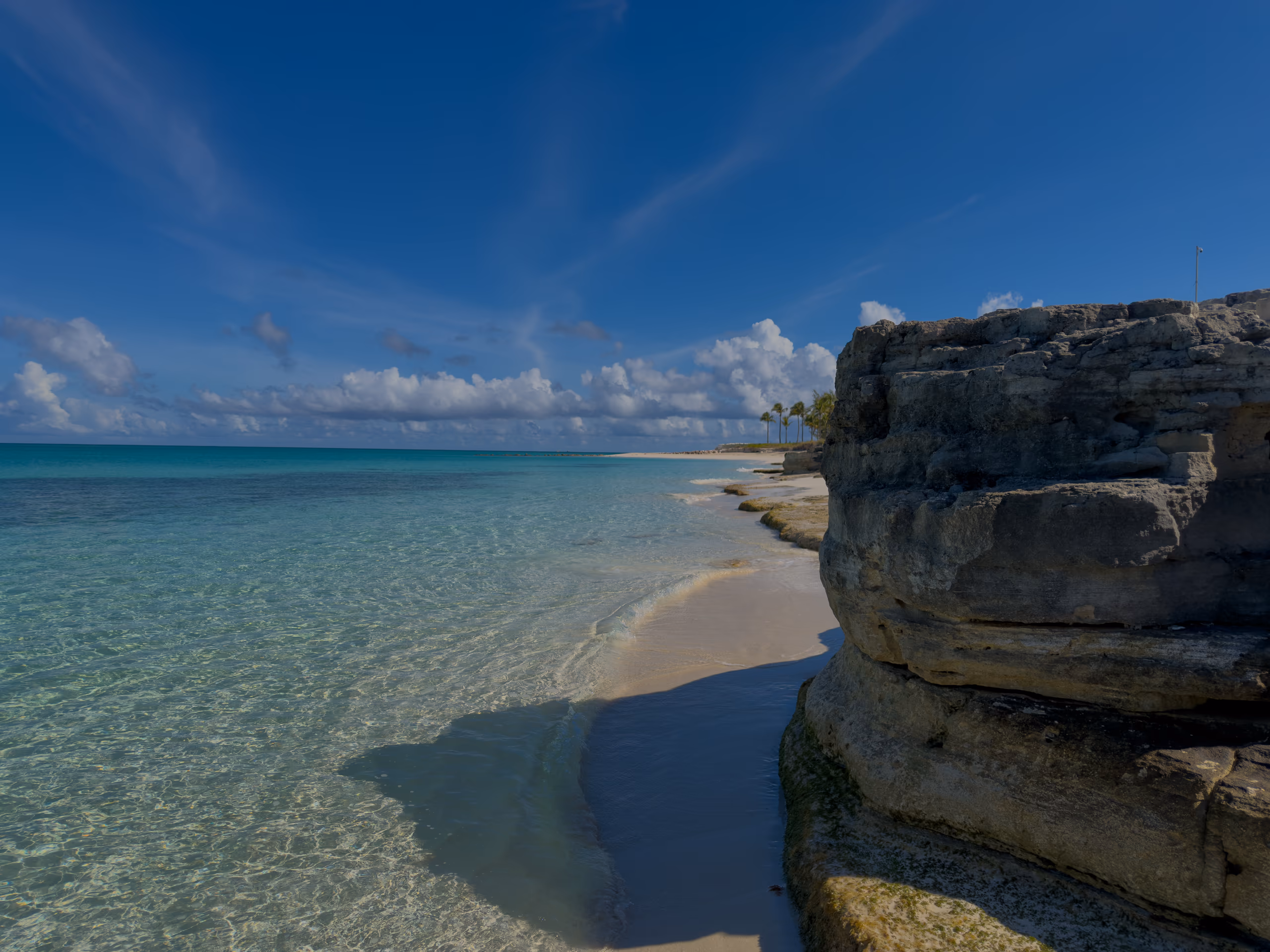Clear turquoise shallows along a white-sand beach, beside a layered limestone rock outcrop