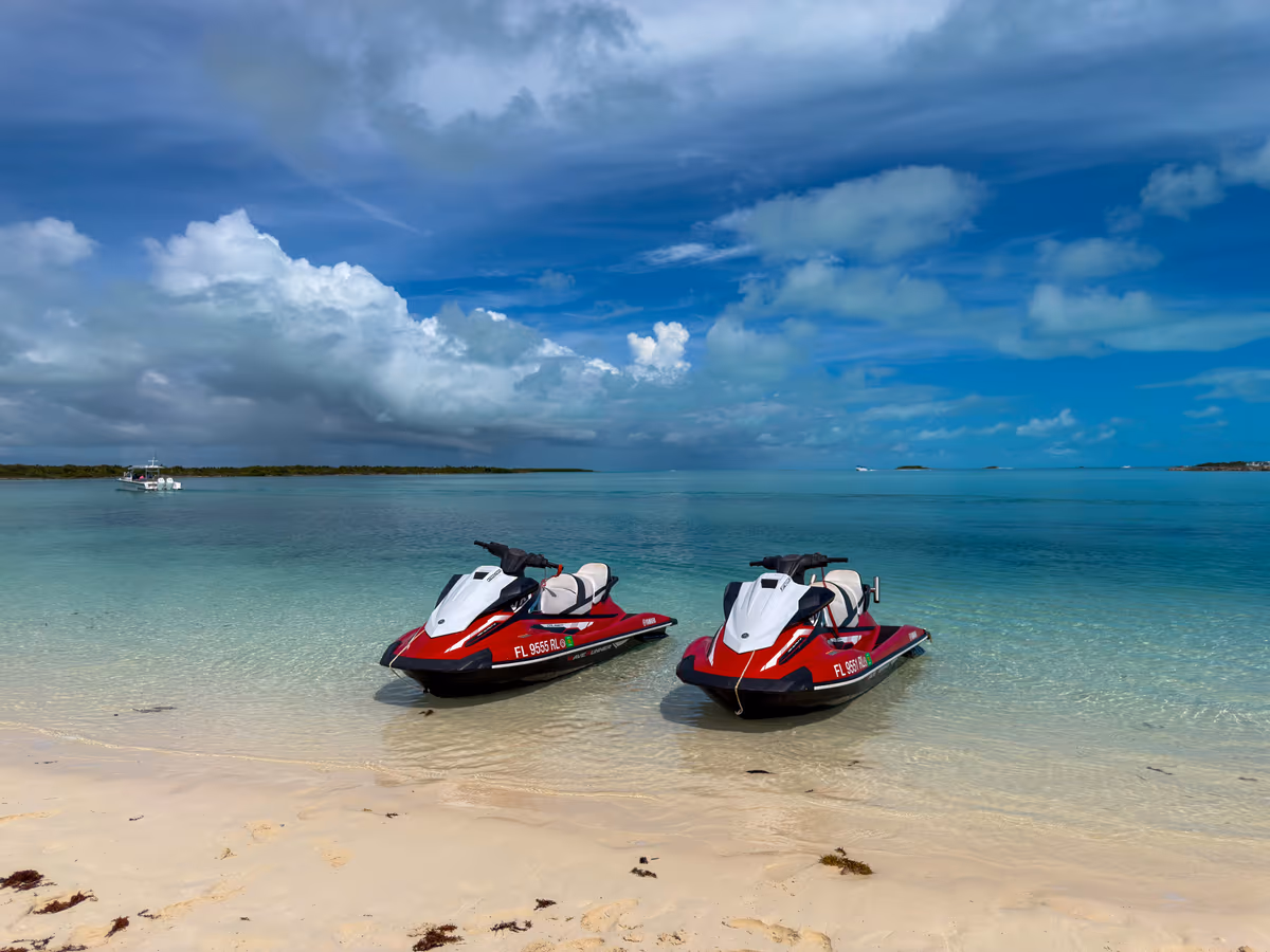 Two red and white Jet Skis beached in clear shallow water on a white sand beach, with a boat and storm clouds on the horizon