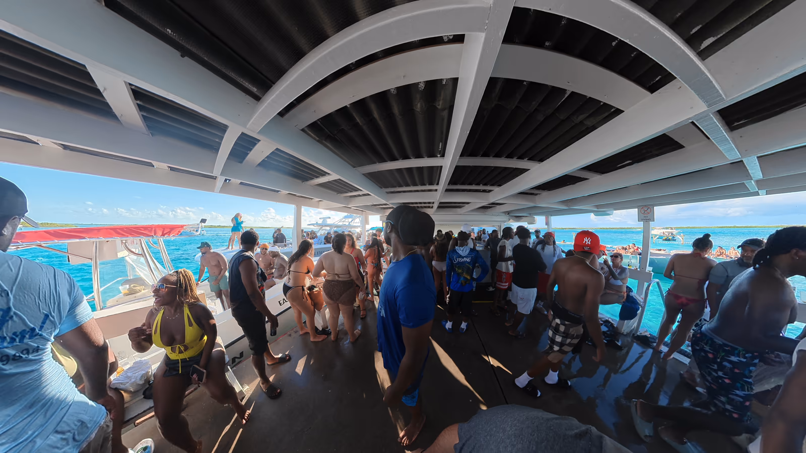 A crowd of guests under the covered deck at Noah's Ark, with turquoise water and moored boats past the railing