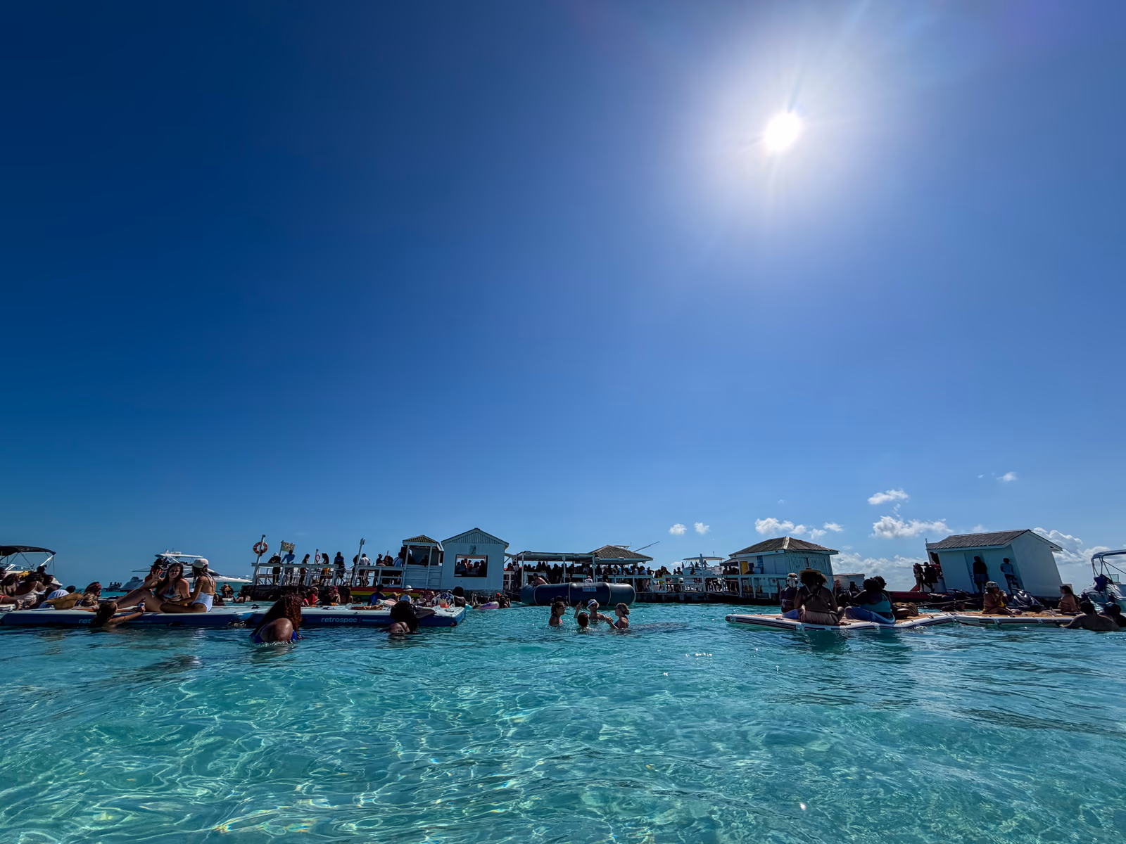 The Noah's Ark floating bar seen from the water, guests standing in the clear turquoise shallows in front of it