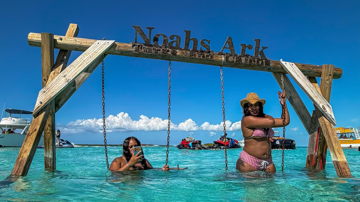 Two guests on the swing set under the Noah's Ark sign, standing in clear turquoise shallows