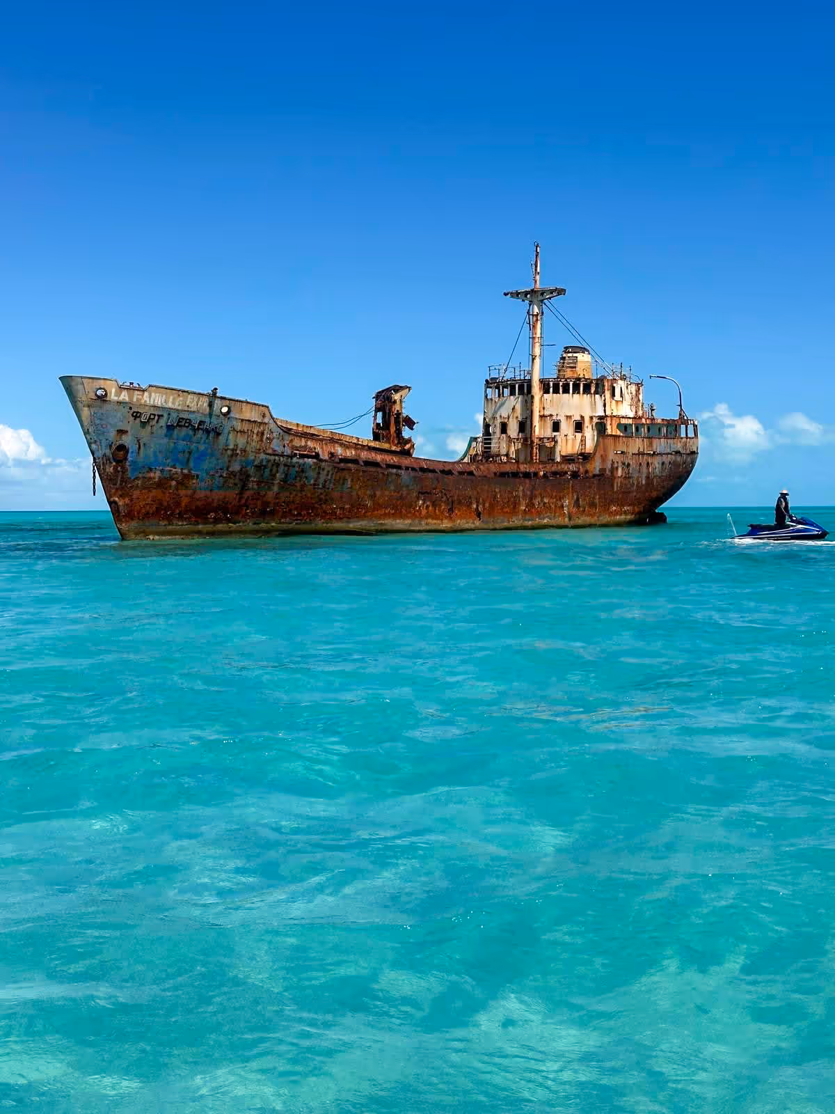 The rusted La Famille Express shipwreck in clear turquoise water, with a guest on a Jet Ski beside its stern