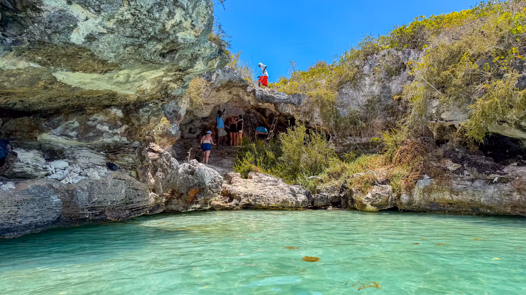 The open mouth of Pirate's Cave in the limestone bluff, visitors inside, clear turquoise water in front