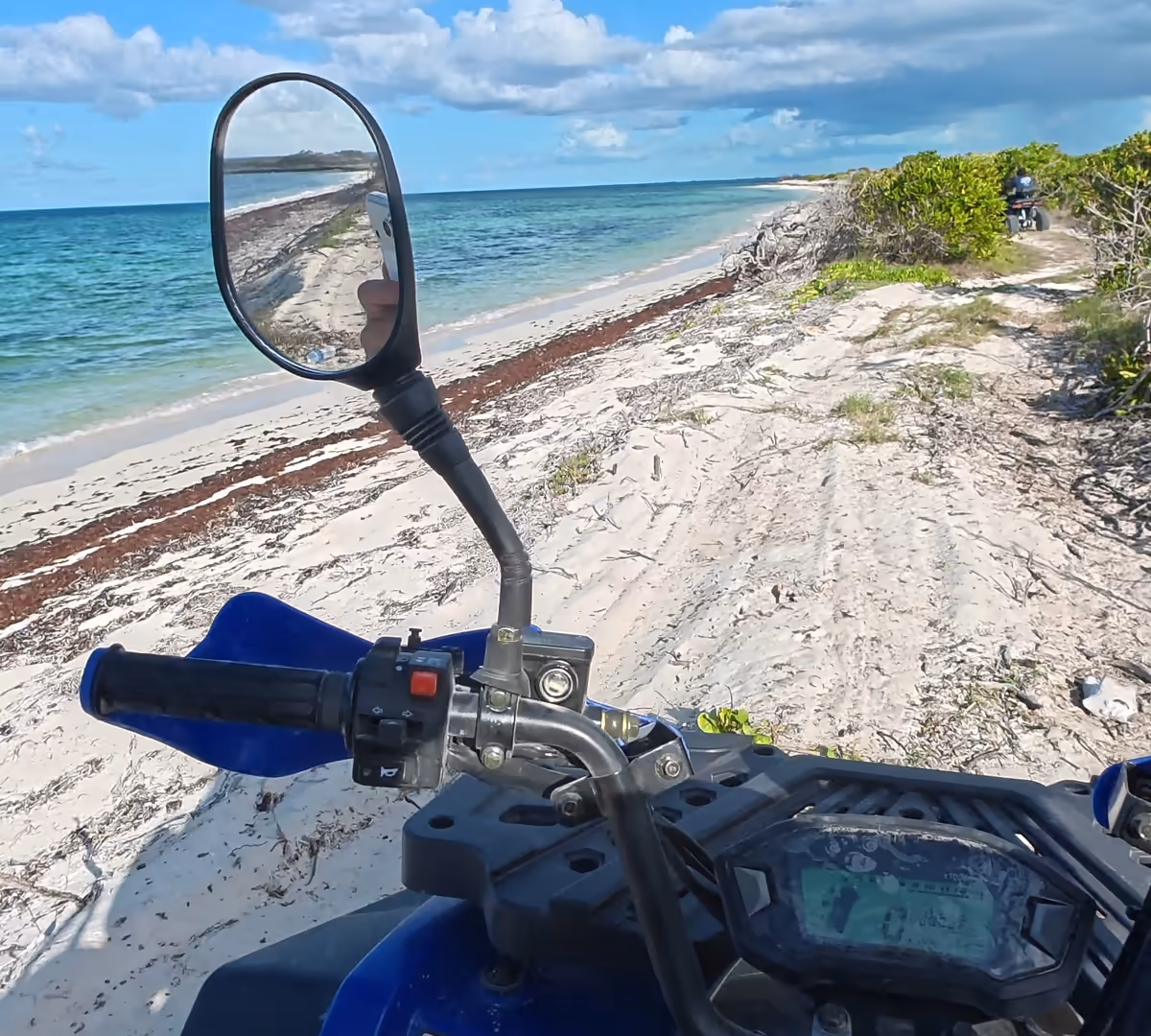 Rider view over the handlebars of a blue quad on a sand trail along the shore, turquoise sea and clouds to the left and another quad ahead
